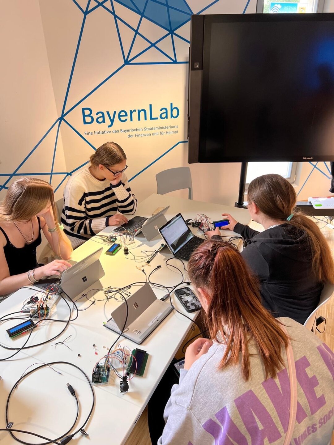 Jugendliche tüfteln und programmieren im Sommercamp Fit4IT 4 junge Frauen sitzen an Laptops an einem Tisch mit vielen Kabeln. Im Hintergrund der Schriftzug